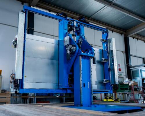 The image shows an industrial system equipped with an automated handling system, characterized by a machine with robotici arms and control panels, used for operations of assembly or processing. The context suggests applications in sector manufacturing, with a focus on automation processes to improve production efficiency.